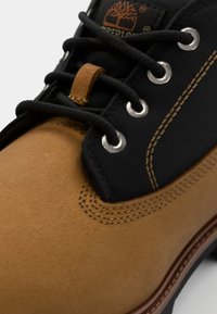 Brune suedeboots med sorte tekstil detaljer, metalløye, kontraststing og en lærpulltab på tungen for enkel påføring.