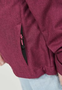 Gros plan sur une personne portant une veste rose foncé texturée avec une poche latérale zippée, et un jean bleu clair visible en bas.