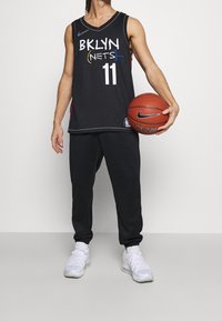Black basketball jersey with "BKLYN NETS" and number "11," featuring herringbone texture. Paired with black sweatpants and white athletic shoes.