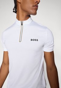 Biela polo košeľa so krátkym zipsom, textúrovaná tkanina s malými perforáciami a čierny nápis "BOSS" na ľavej hrudi.