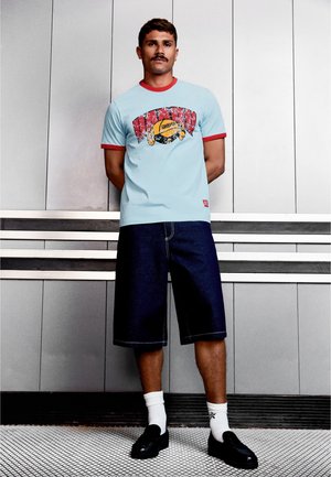 Mężczyzna z wąsami, ubrany w jasnoniebieski T-shirt z grafiką, ciemne krótkie spodenki do kolan, białe skarpetki i czarne buty, stojący przy ścianie z paneli.