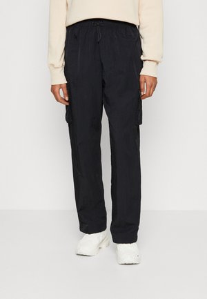 Person trägt schwarze Cargo-Hosen mit Kordelzug in der Taille und Seitentaschen, kombiniert mit weißen klobigen Sneakern und einem cremefarbenen Sweatshirt.
