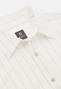 Camicia bianca con sottili strisce nere, colletto strutturato e apertura con due bottoni. Il tessuto sembra liscio con una leggera trama. Etichetta del marchio visibile.