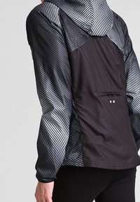 Giacca con cappuccio nera e grigia con strisce diagonali, dotata di una tasca posteriore con zipper e realizzata in un materiale leggero e resistente all'acqua.