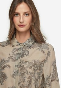 Beige skjorta med knäppning framtill som har ett paisleymönster i bruna och silverfärger, med klassisk krage och en mjuk tygtextur.