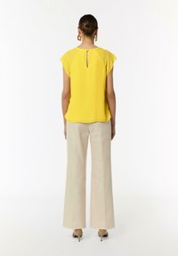 Żółta bluzka z krótkimi rękawami i detalem w kształcie klucza na plecach, zestawiona z beżowymi spodniami o szerokich nogawkach, zaprezentowana na neutralnym tle.