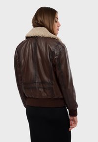 Blouson en cuir brun avec un col en fourrure beige, des poignets et un ourlet côtelés, présentant des coutures visibles et un design court.