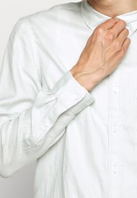 Ljusgrön linnekostym med krage som kan knäppas, långa ärmar och vita knappar. Texturen verkar något rynkad och luftig.