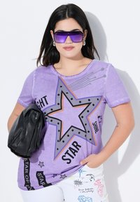 Camiseta corta de manga en color púrpura con un gráfico de estrella, lunares negros y acentos de texto. Combinada con un bolso negro y pantalones blancos con estampado.