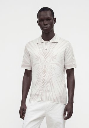 Bărbat purtând un tricou polo cu mâneci scurte, de culoare deschisă, cu un model geometric subtil și pantaloni albi, pe un fundal simplu.
