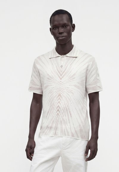 Homme portant un polo à manches courtes, de couleur claire, avec un motif géométrique subtil et un pantalon blanc sur un fond uni.