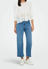 Weiße bestickte Bluse mit Puffärmeln und gewelltem Saum, kombiniert mit hochgeschnittenen blauen Jeans und weißen Sneakers.