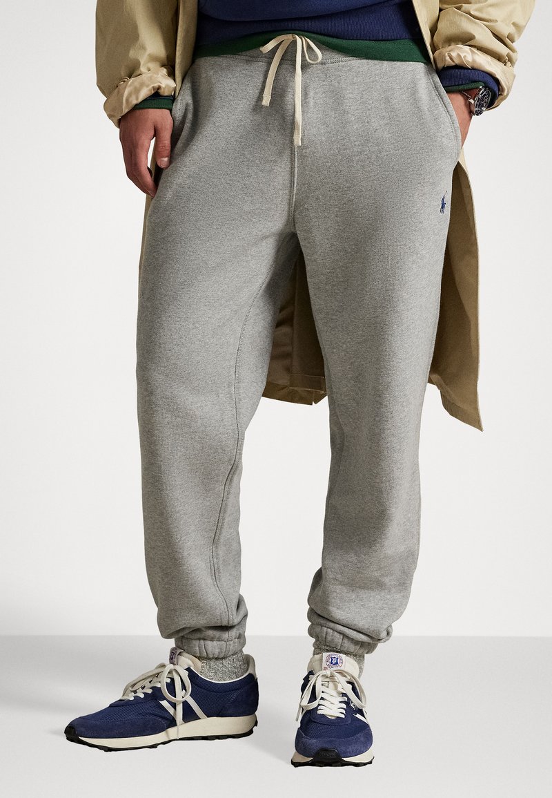 Pantaloni de trening gri din jerseu, cu talie elastică și mansete, prevăzuți cu buzunare laterale și un mic logo albastru. Asortați cu pantofi de sport albastre.