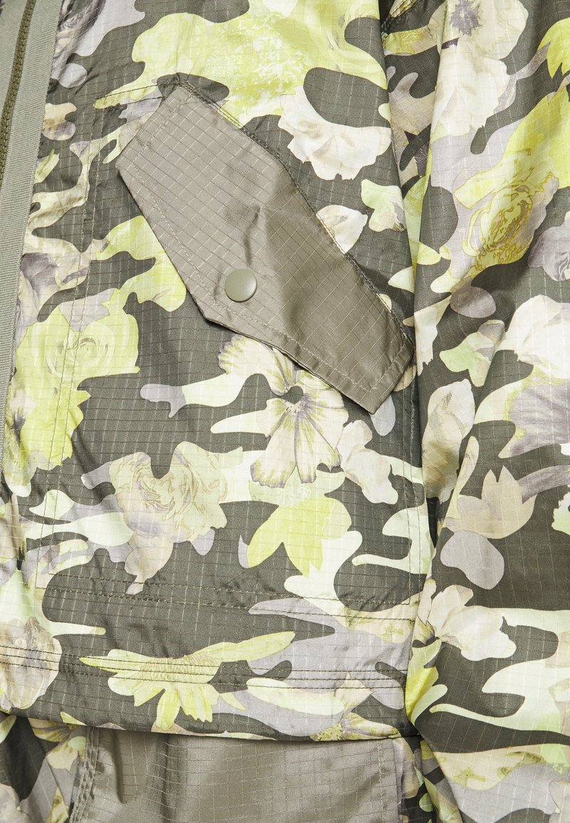 Chaqueta de camuflaje en tonos verdes y amarillos, con una textura brillante y un bolsillo con solapa que cuenta con un cierre a presión.