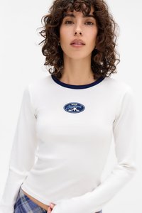 Młoda kobieta z kręconymi włosami nosi biały longsleeve z granatowym kołnierzykiem i owalną, vintage naszywką na piersi, w zestawieniu z niebieskimi spodniami w kratę.
