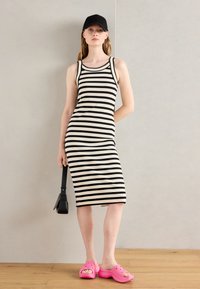Robe midi à rayures noires et blanches avec une silhouette ajustée, portée avec des sabots roses et un sac noir. L'arrière-plan est neutre.
