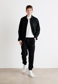 Zwarte blouse over een witte T-shirt met ronde hals, gecombineerd met zwarte sweatpants met een logo, en witte sneakers. Neutrale achtergrond.