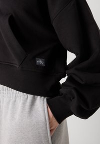 Čierna mikina s rebrovanými manžetami a lemom, so prednou vreckom a štítkom s logom Calvin Klein. V kombinácii so sivými teplákmi.