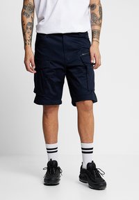 Cargo shorts in navy blue fabric, featuring multiple pockets, a zip pocket detail, and a relaxed fit. Worn with white socks and black sneakers.