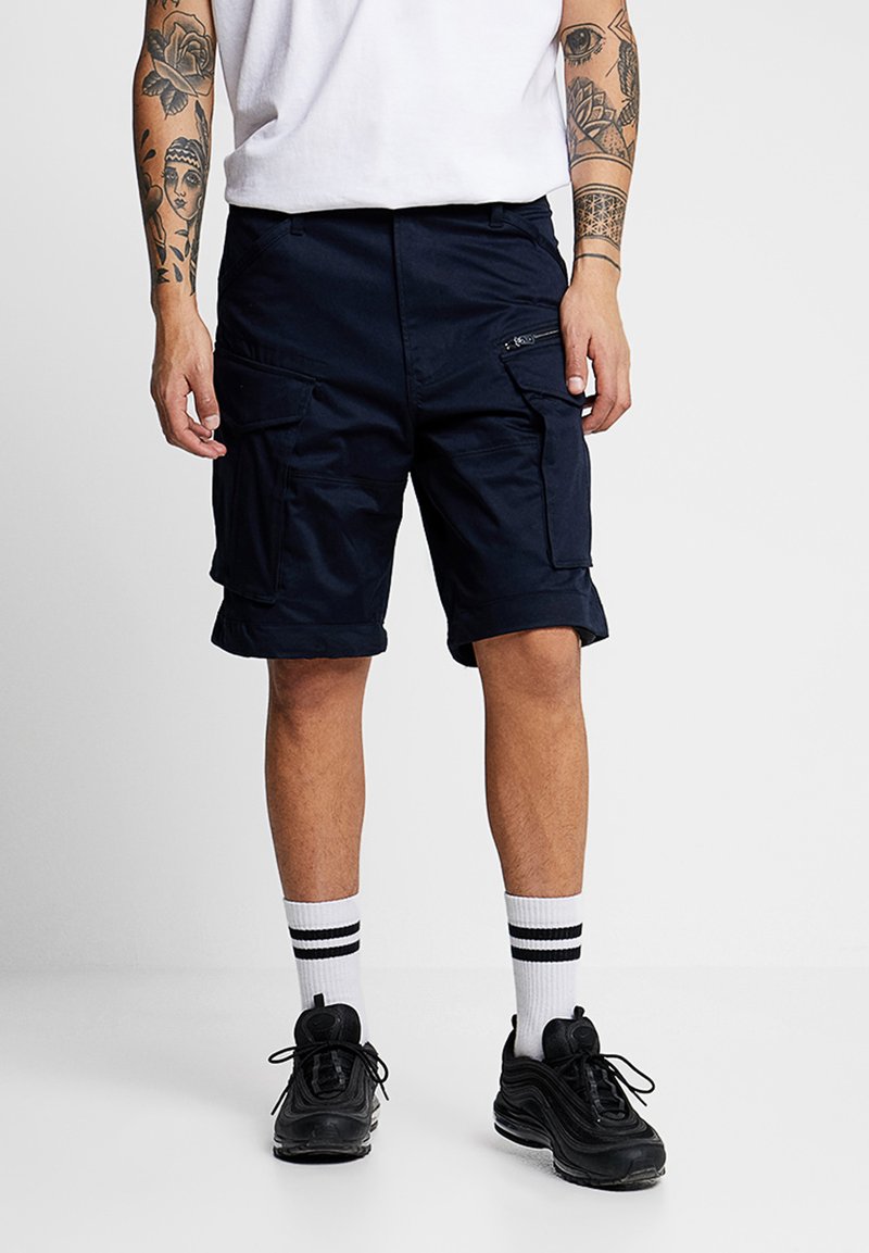 Cargo shorts in navy blue fabric, featuring multiple pockets, a zip pocket detail, and a relaxed fit. Worn with white socks and black sneakers.