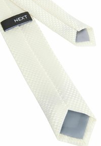 Cravate blanche texturée avec un motif en losanges, présentant un dessous gris et une étiquette noire "NEXT". Extrémités triangulaires pointues.