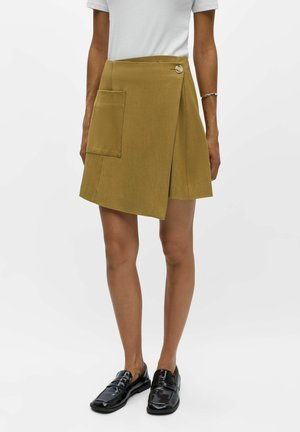 Femme portant une jupe portefeuille jaune moutarde avec une grande poche et un bouton, un haut blanc, un bracelet argenté et des mocassins noirs vernis.