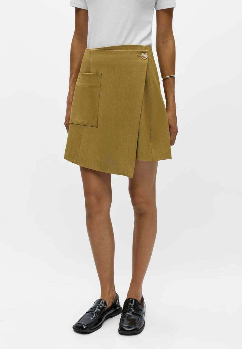 Femme portant une jupe portefeuille jaune moutarde avec une grande poche et un bouton, un haut blanc, un bracelet argenté et des mocassins noirs vernis.