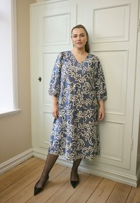 Modrá kvetinová šaty s výstrihom do V, trojštvrťovými rukávmi a voľným strihom. Nosené s čiernymi podpätkami a priesvitnými pančuchami.