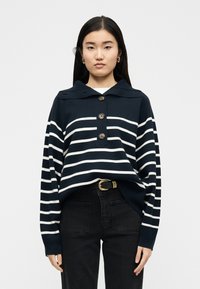 Femme portant un pull à rayures bleu marine et blanc avec trois boutons et un pantalon noir avec une ceinture à boucle dorée, debout devant un fond uni.