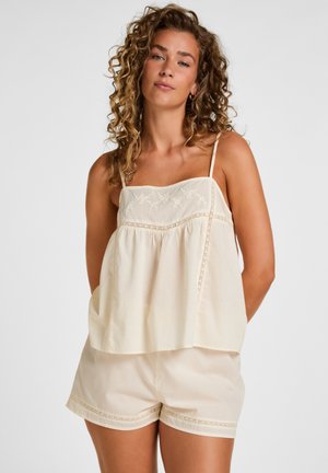 Jeune femme aux cheveux bouclés portant un haut sans manches crème avec broderies et un short assorti sur un fond uni.