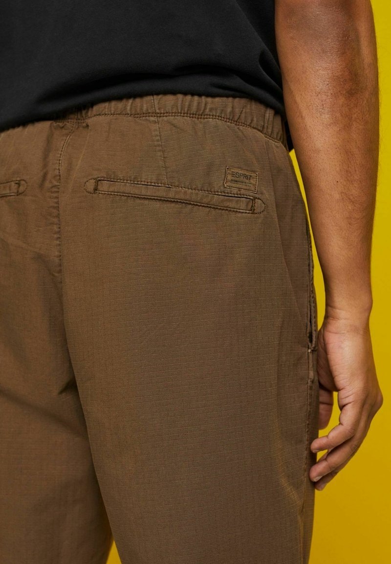 Homme portant un pantalon décontracté marron avec une ceinture élastique et des poches passepoilées à l'arrière, debout devant un fond jaune.
