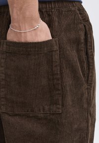 Brown corduroy pants with a vertical ribbed texture, featuring a large side pocket and a fitted waistband. Silver chain bracelet on wrist.