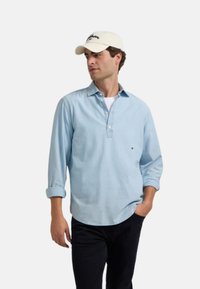 Camisa de manga larga azul claro con cuello abotonado y puños remangados, combinada con una camiseta blanca y pantalones negros. Gorro crema expuesto.