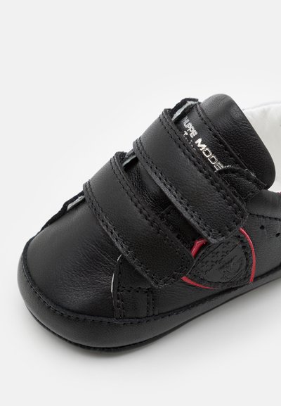 Sapatos de bebé em couro preto com duas tiras de fecho de velcro, biqueira arredondada, costura decorativa e um detalhe em vermelho. Forro interno suave visível.
