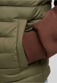 Polstret olivengrønn vest med en sidelomme og en brun ribbestrikket mansjett. Materialet har en glatt tekstur med quiltede detaljer.