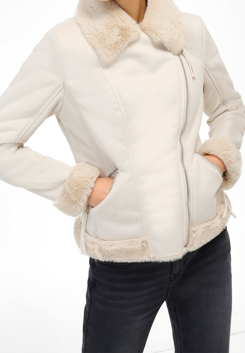 Femme portant une veste crème avec col, poignets et ourlet en fourrure douce, mains dans les poches zippées, associée à un pantalon noir.