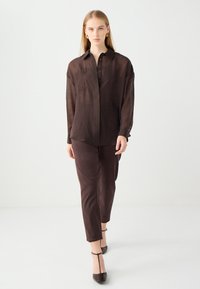 Camicia marrone con bottoni e design ricamato, abbinata a pantaloni marroni coordinati. Tessuto morbido e leggero; vestibilità su misura e leggera lucentezza.