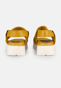 Gelbe Leder-Sandalen mit breiten Riemen, offenem Rücken und einer klobigen weißen Sohle; glatte Textur mit minimaler Gestaltung.