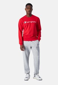 Sudadera roja con el logo blanco de "Champion", combinada con pantalones de chándal gris claro. Modelo viste zapatillas blancas y está de pie frente a un fondo sencillo.
