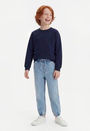 Ragazzo dai capelli rossi che sorride, indossa una felpa blu navy, jeans con vita elasticizzata azzurri e scarpe da ginnastica bianche, in piedi davanti a uno sfondo bianco uniforme.