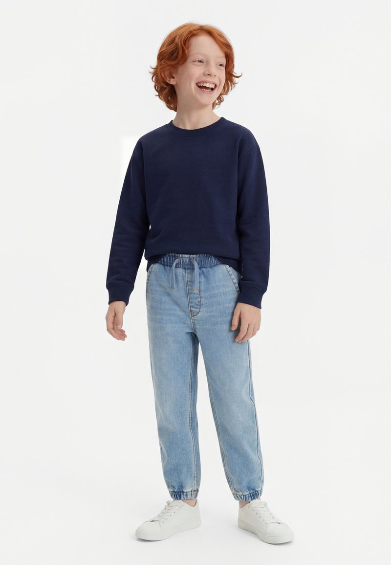 Κοκκινομάλλης αγόρι που χαμογελά, φοράει μπλε σκούρο φούτερ, τζιν με ελαστική μέση ανοιχτού γαλάζιου χρώματος και λευκά αθλητικά παπούτσια, στέκεται μπροστά σε απλό λευκό φόντο.