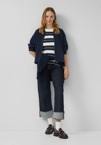 Marine bomberjack over een wit met marineblauw gestreept shirt, gecombineerd met donkere wijde spijkerbroek met denim cuffs, en stevige bruine loafers.