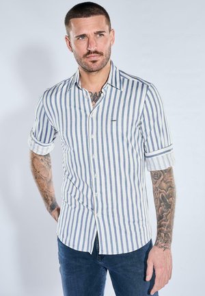 Mann mit kurzen Haaren und Tattoos, der ein weißes Hemd mit blauen vertikalen Streifen und hochgekrempelten Ärmeln trägt, kombiniert mit dunkelblauen Jeans.