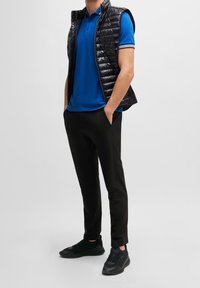 Mężczyzna w niebieskiej koszulce polo, czarnej pikowanej kamizelce bez rękawów, czarnych spodniach i czarnych trampkach, stojący z rękami w kieszeniach.