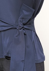 Tissu en satin bleu marine noué en un nœud autour de la taille sur un top bleu marine assorti et un pantalon noir.
