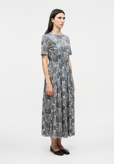 Femme debout, portant une robe midi à motifs floraux bleus et blancs avec des manches courtes et des ballerines noires, sur un fond uni.