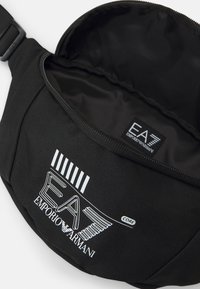 Juodas audinys juosmens krepšys su užtrauktuku. Pabrėžia baltą „EA7 Emporio Armani“ logotipą ir vertikalias juosteles. Raižyta medžiaga. Vidaus paviršius lygus.