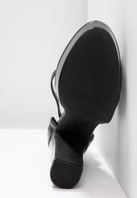 Chaussure à talon haut noire montrée semelle vers le haut, positionnée contre un coin blanc avec semelle texturée et talon carré.