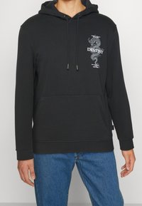 Sudadera negra con bolsillo frontal, gráfico de dragón blanco y la palabra "DESTINO" en el pecho, combinada con jeans azules.