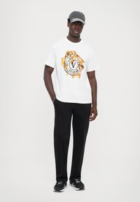 Hvit T-skjorte med fargerik grafisk logo, svarte straight-leg bukser, svarte joggesko og grå caps med trykket tekst. Minimalistisk bakgrunn.